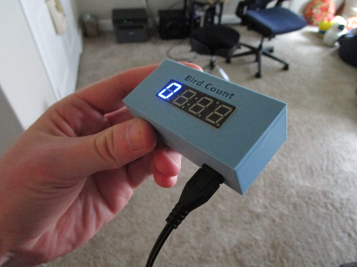 A hand holding the 3D-printed indoor counter unit, its blue 7-segment display reading the current bird count